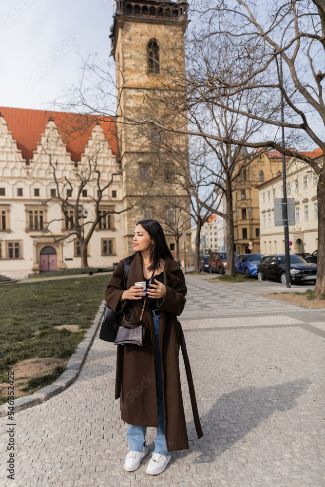 Trendy young woman in coat holding coffee to go on urban street in Prague.