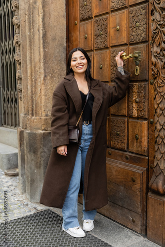 happy brunette woman in trendy coat touching door handle near building in prague. photo