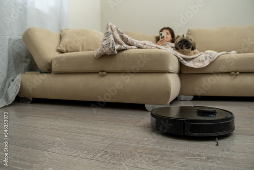 Black robotic vacuum cleaner works in light living room. Covered with warm blanket woman rests lying near shih tzu on beige sofa looking at phone