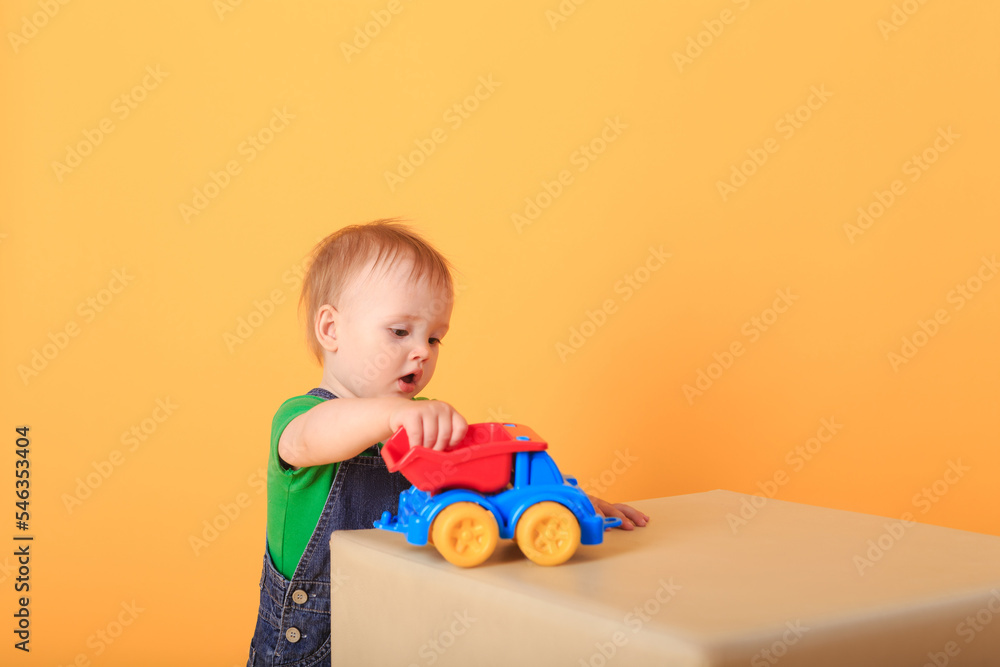 A little handsome boy plays with a toy on a light-colored pouffe . Studio shot of a baby boy against a yellow background. A little handsome boy plays with a toy on a light-colored pouffe . Studio shot of a baby boy against a yellow background.