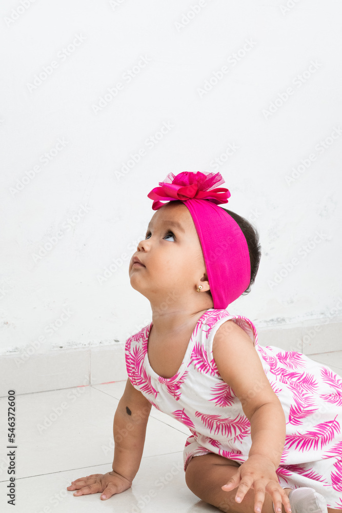 baby latina sitting on the floor looking upwards, with a bench dress with fuchsia leaves. white background baby latina sitting on the floor looking upwards, with a bench dress with fuchsia leaves. white background
