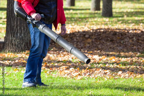 Worker operating heavy duty leaf blower in city park. Removing fallen leaves in autumn. Park cleaning service