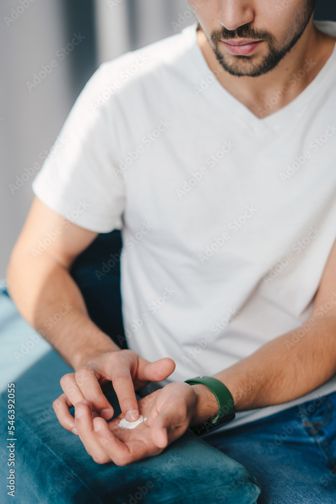 Close-up portrait of a man applying cream onto hand on sofa. Beauty treatment. Dermatology, cosmetology. Perfect cosmetology skin care. Body care. Close-up portrait of a man applying cream onto hand on sofa. Beauty treatment. Dermatology, cosmetology. Perfect cosmetology skin care. Body care.