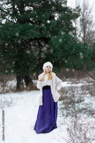Bright color photo of a beautiful young lady dressed in vintage clothes in a fur hat and vest in a long purple skirt in a winter coniferous forest with pines