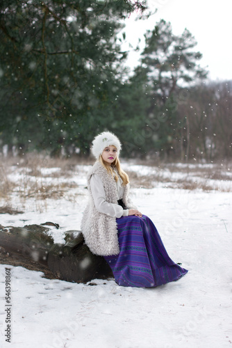 Bright color photo of a beautiful young lady dressed in vintage clothes in a fur hat and vest in a long purple skirt in a winter coniferous forest with pines