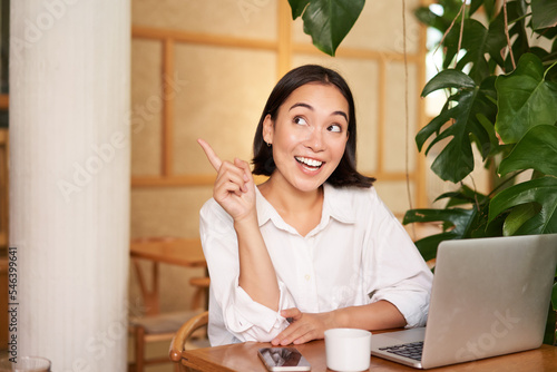 Stylish young entrepreneur, girl with laptop sitting in cafe, pointing finger at advertisement banner on upper left corner photo