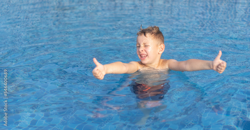 Little blond boy in the outdoor pool is showing thumbs up