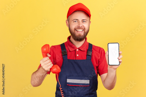 Portrait of delighted smiling worker man holding out retro phone and cell phone with empty screen for advertisement, choose best device for you, Indoor studio shot isolated on yellow background. photo