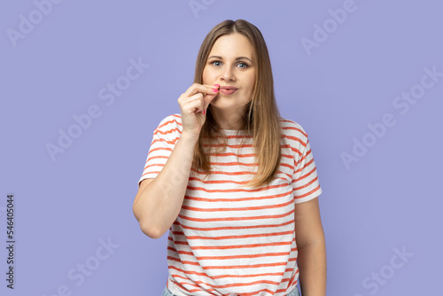 Portrait of beautiful positive blond woman wearing striped T-shirt standing and zipping her mouth, knows secret, does not tell to anyone. Indoor studio shot isolated on purple background.
