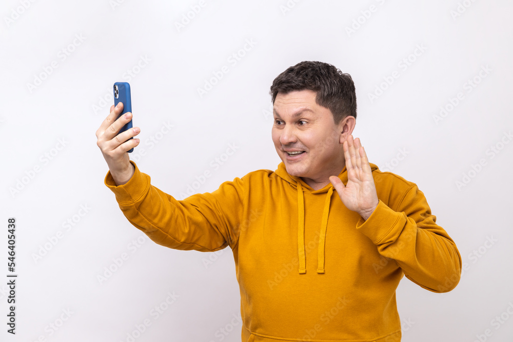 Cheerful middle aged man saying hi making video call using smartphone, smiling with toothy smile, 4g connection, wearing urban style hoodie. Indoor studio shot isolated on white background. Cheerful middle aged man saying hi making video call using smartphone, smiling with toothy smile, 4g connection, wearing urban style hoodie. Indoor studio shot isolated on white background.