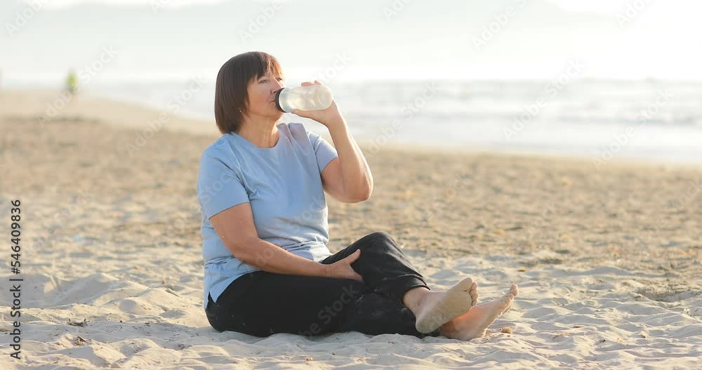 Adult middle aged woman is drinking water after run, yoga, sport exercises on beach by the sea and enjoying early morning with beautiful sunrise on summer day. People do sports, running on background. Adult middle aged woman is drinking water after run, yoga, sport exercises on beach by the sea and enjoying early morning with beautiful sunrise on summer day. People do sports, running on background.