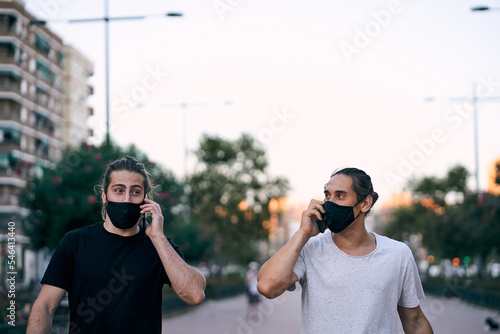 Fototapeta two caucasian young men walking on the city boulevard with face mask talking on