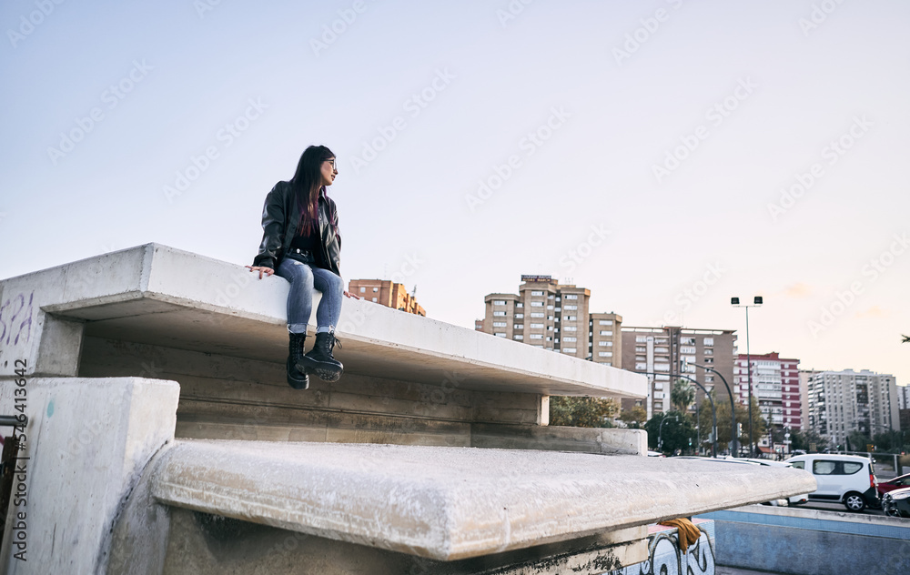 latina girl in ripped blue pants fur jacket glasses looking at the city sitting calm and relaxed on the concrete wall latina girl in ripped blue pants fur jacket glasses looking at the city sitting calm and relaxed on the concrete wall
