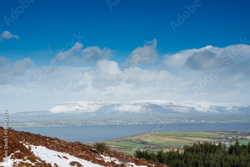 Hill with red grass and green forest. Benbulben flat top mountain covered with snow and cloudy sky in the background. County Sligo, Cold winter season in Ireland. Irish landscape.