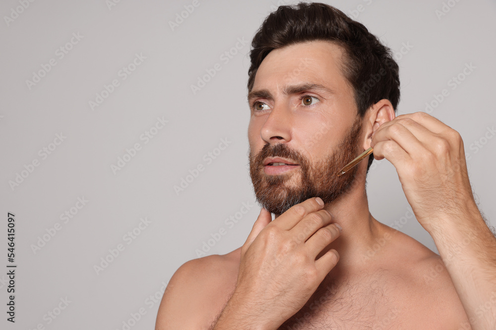 Obraz premium Man applying oil onto beard on grey background. Space for text