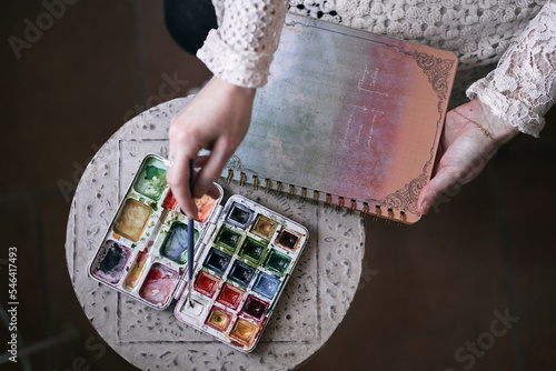 closeup of a case of watercolors of many colors and a hand holding a fine brush ready to paint on a notebook