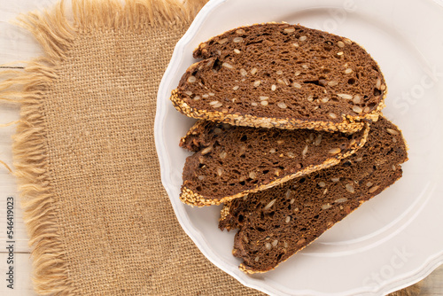 Several pieces of rye multigrain bread with a white plate and jute napkin on a wooden table, macro, top view.
