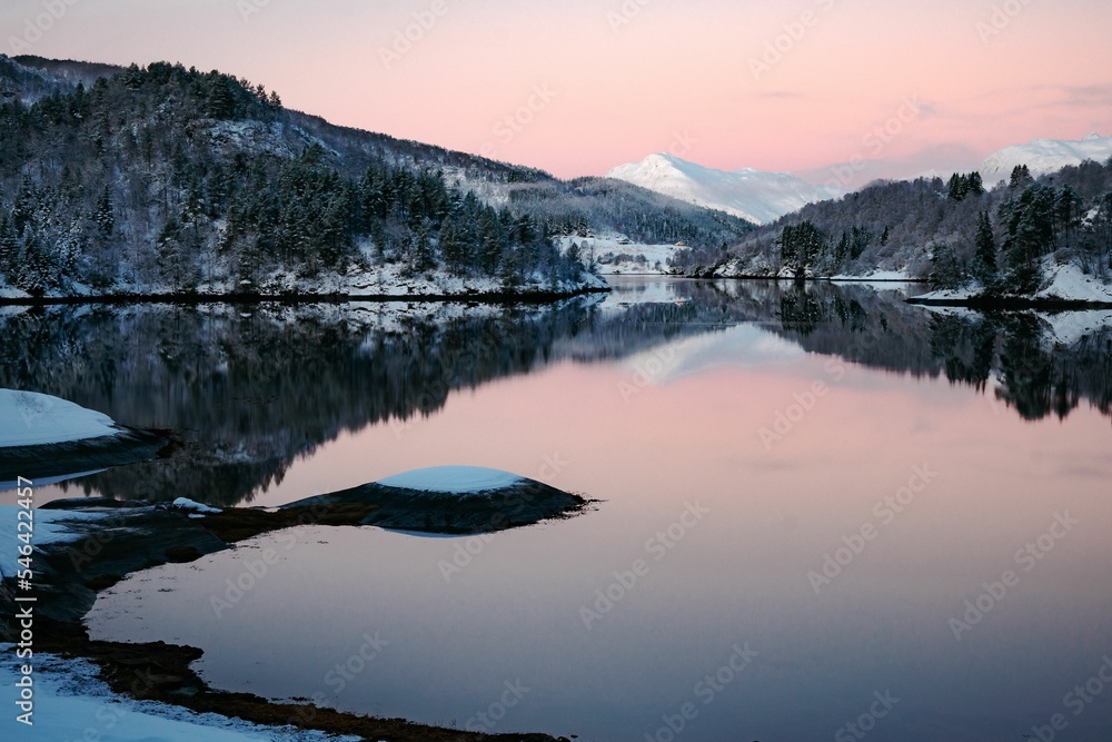 Naklejka premium Beautiful view of a lake surrounded by tree-covered snowy mountains under a pink cloudy sky