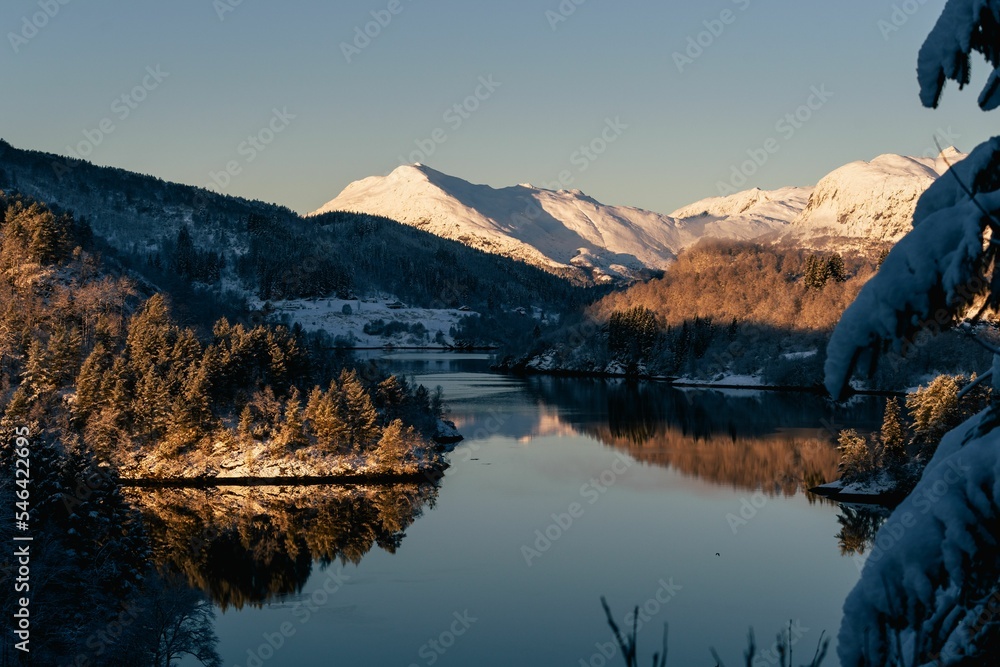 Naklejka premium Beautiful view of a lake surrounded by tree-covered snowy mountains under a clear blue sky