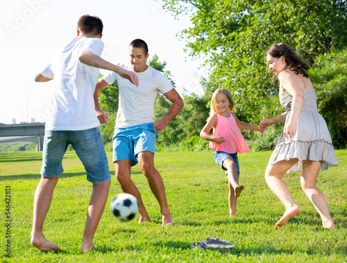 Happy friendly family with children playing football on green lawn in summer city park