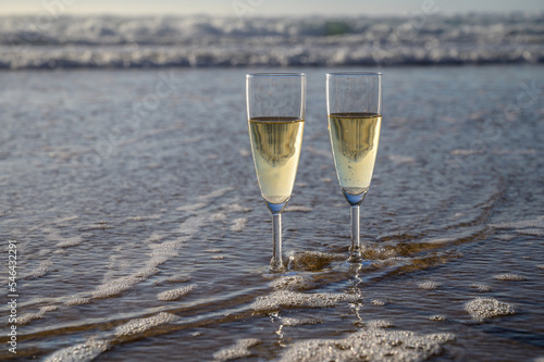 Glasses of cava or champagne sparkling wine on white sandy ocean beach with water waves on sunset in sunlights