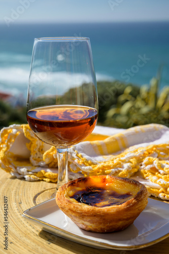 Portugal's traditional food and drink, glass of porto wine or muscatel and sweet dessert Pastel de nata egg custard tart pastry served with view on blue Atlantic ocean in Lisbon area, Portugal photo