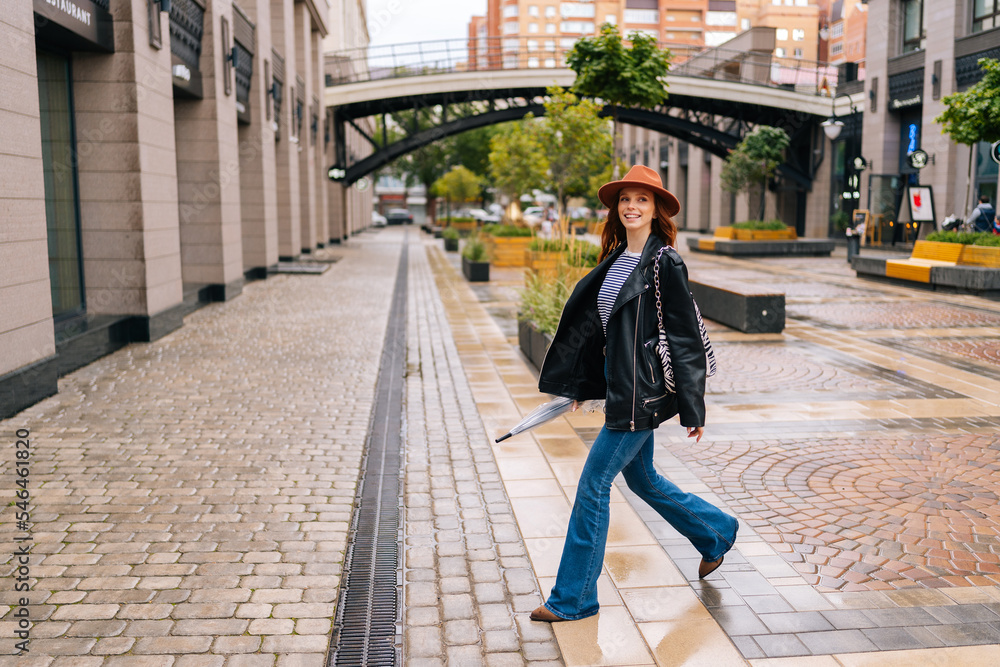Full length of positive redhead woman wearing fashion hat walking on European city street holding in hand closed transparent umbrella. Concept of modern female lifestyle at autumn season.