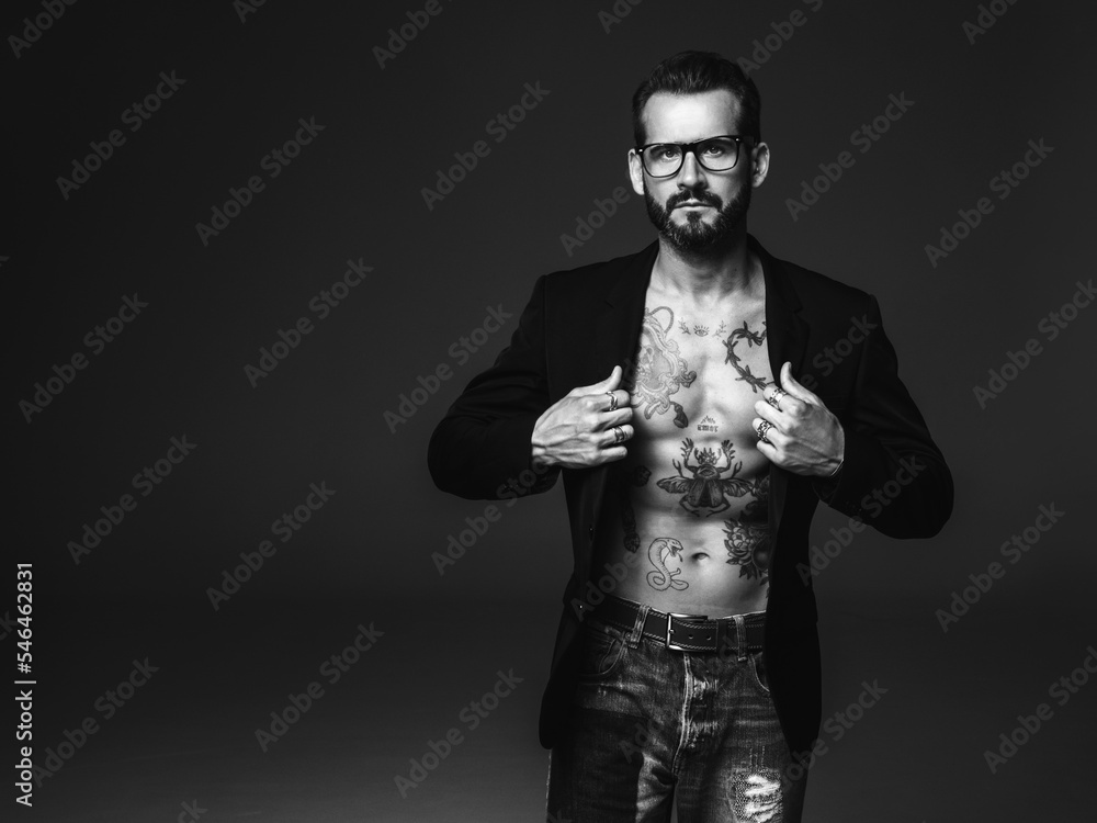 Portrait of handsome confident stylish hipster lambersexual model. Sexy modern man dressed in black jacket. Naked torso with tattoos.Fashion male posing in studio on dark background. In spectacles Portrait of handsome confident stylish hipster lambersexual model. Sexy modern man dressed in black jacket. Naked torso with tattoos.Fashion male posing in studio on dark background. In spectacles