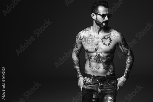 Portrait of handsome confident stylish hipster lambersexual model. Sexy modern man. Naked torso with tattoos.Fashion male posing in studio on dark background. In sunglasses © halayalex