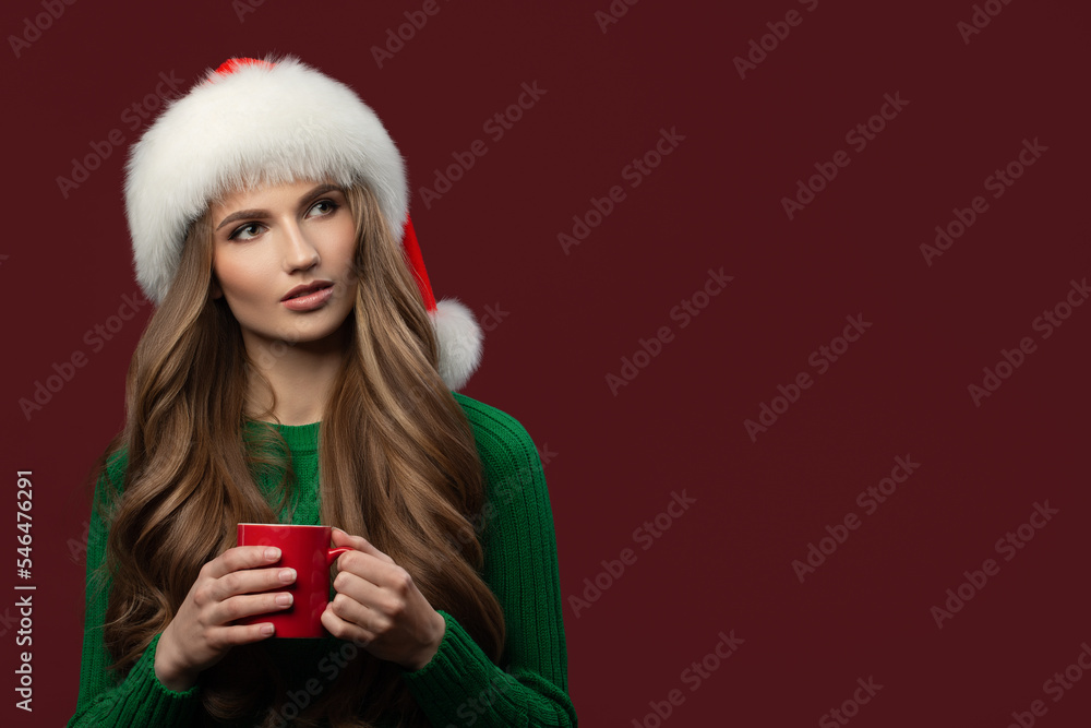 Portrait of a girl in a santa hat. Holds a mug of coffee in her hand. Long curly hair. Burgundy background. Portrait of a girl in a santa hat. Holds a mug of coffee in her hand. Long curly hair. Burgundy background.