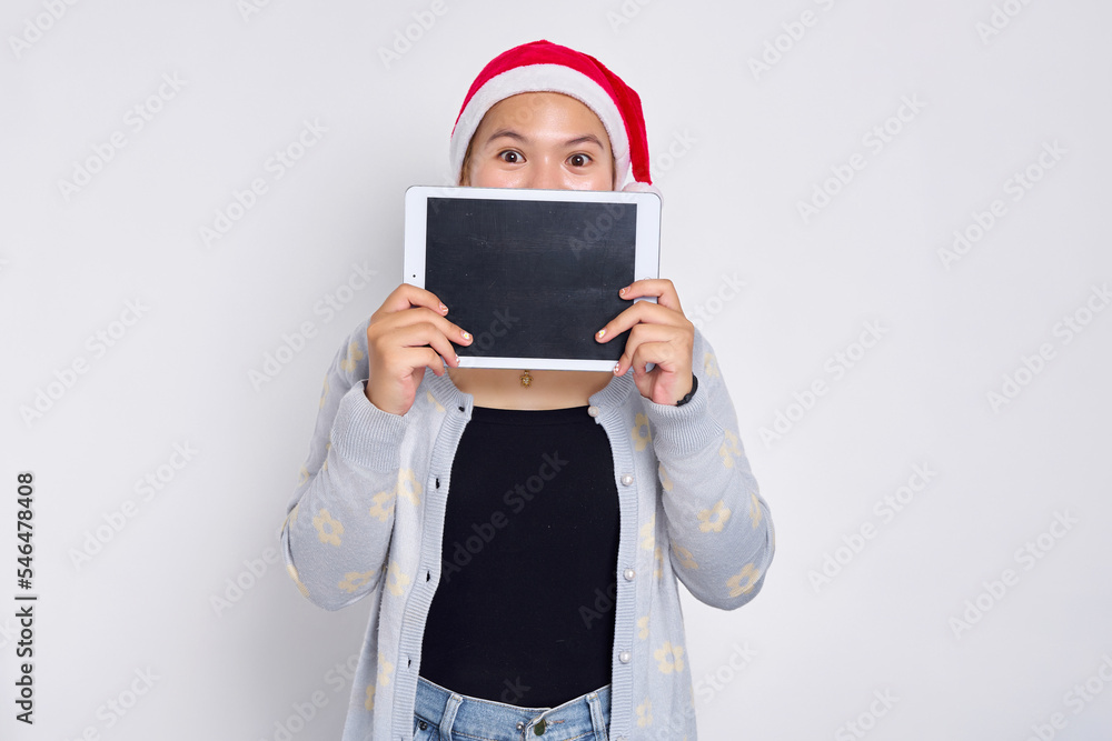 young Asian woman in a hat Christmas covering her face on blank screen tablet isolated over white background. Indonesian people celebrate Christmas concept