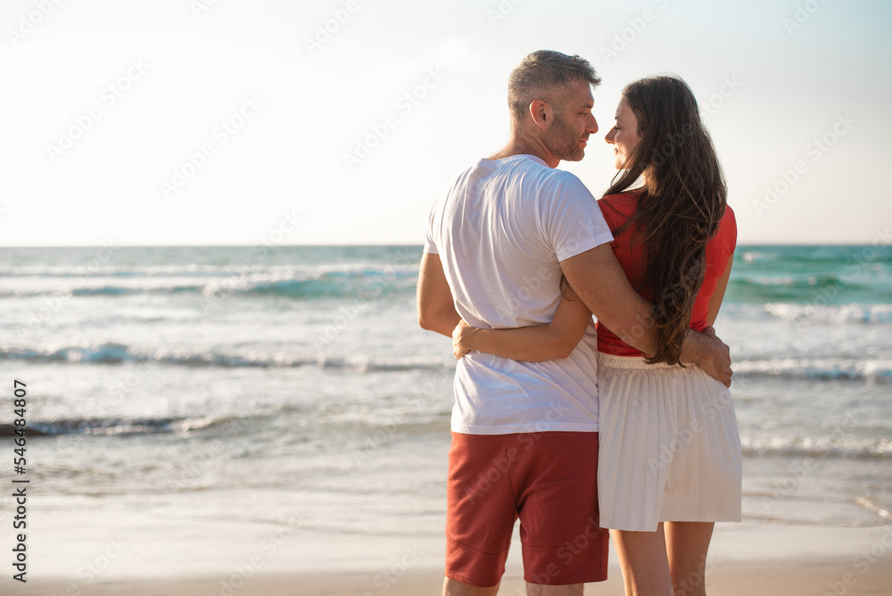 Happy couple hugs on beach on background clear sky and blue sea. Romantic date on seaside. Good weather and calm water surface. White and red clothes. Back view. Happy couple hugs on beach on background clear sky and blue sea. Romantic date on seaside. Good weather and calm water surface. White and red clothes. Back view.