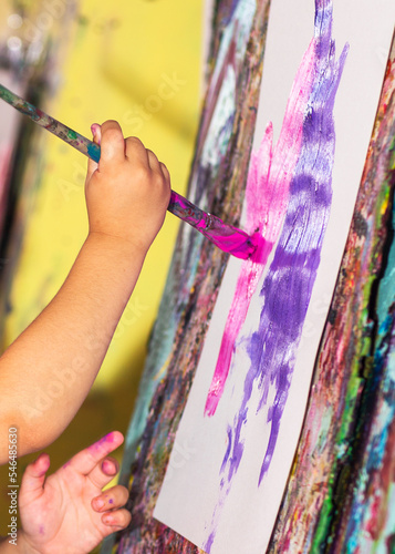 Kid's hand holds paintbrush. A kid paints with brush on a wight paper. 