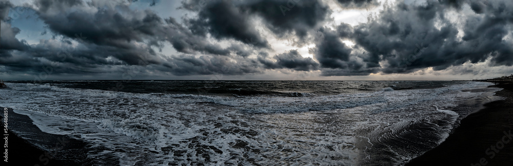 Scary weather sunset panoramic on the beach with dramatic sky with dark cumulus clouds and stormy sea with waves breaking on the shore generating white foam