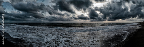 Scary weather sunset panoramic on the beach with dramatic sky with dark cumulus clouds and stormy sea with waves breaking on the shore generating white foam