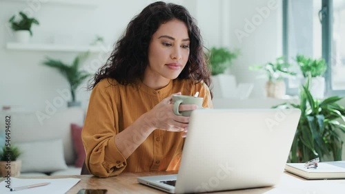 Video of beautiful business woman working with laptop while drinking coffee in living room at home.