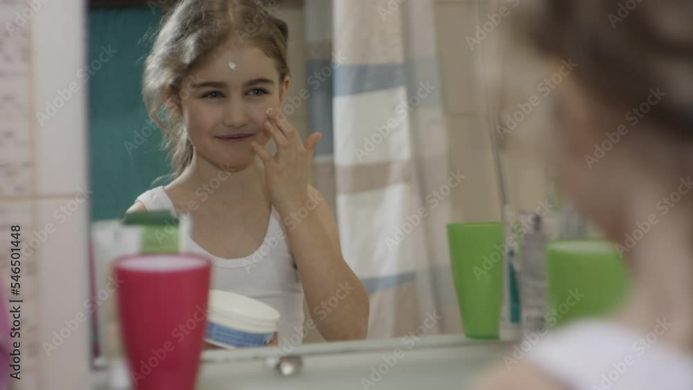 Portrait Young Funny Little Girl Applying Moisturizing Cream on Her Face in Bathroom Looking at Mirror Reflection. Happy Cute Caucasian Toothless Child Applying on Face Cream For Skin Car. Portrait Young Funny Little Girl Applying Moisturizing Cream on Her Face in Bathroom Looking at Mirror Reflection. Happy Cute Caucasian Toothless Child Applying on Face Cream For Skin Car.