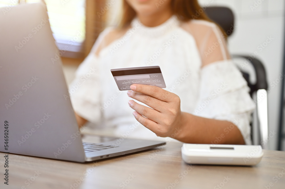 Asian businesswoman sits at her desk holding her credit card and typing on her laptop keyboard Asian businesswoman sits at her desk holding her credit card and typing on her laptop keyboard