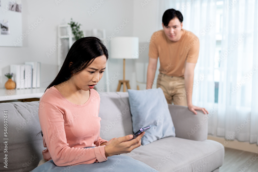 Shocked jealous Asian girlfriend suspecting infidelity by reading text messages on her cheating boyfriend's phone at home. Wife checking husband's smartphone and having jealousy problem Shocked jealous Asian girlfriend suspecting infidelity by reading text messages on her cheating boyfriend's phone at home. Wife checking husband's smartphone and having jealousy problem