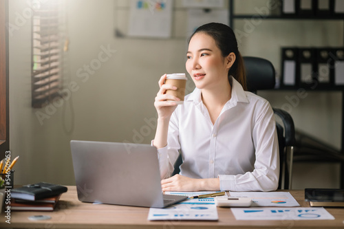 Confident beautiful Asian businesswoman typing laptop computer and digital tablet while holding coffee at office.