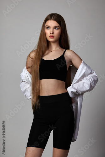 a slender teenager girl in a male oversized white, sports bust, shirt and black bicycle shorts poses in the studio on a dark background