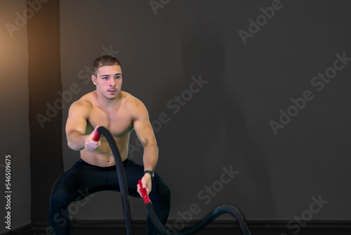 A handsome muscular young man using battle ropes for exercise in a gym. He focuses on performing the exercise. Motivational photo. Copy space.