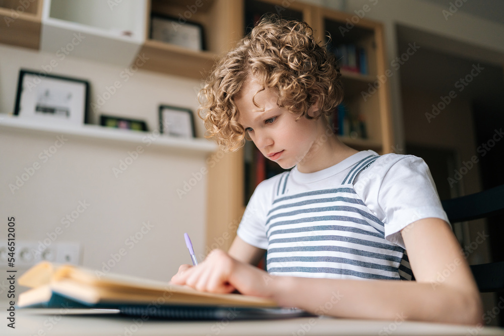 Low-angle view of concentrated little child schoolgirl writing in exercise book doing some homework, sitting at desk near window. Cute curly small school girl studying alone making notes at home. Low-angle view of concentrated little child schoolgirl writing in exercise book doing some homework, sitting at desk near window. Cute curly small school girl studying alone making notes at home.