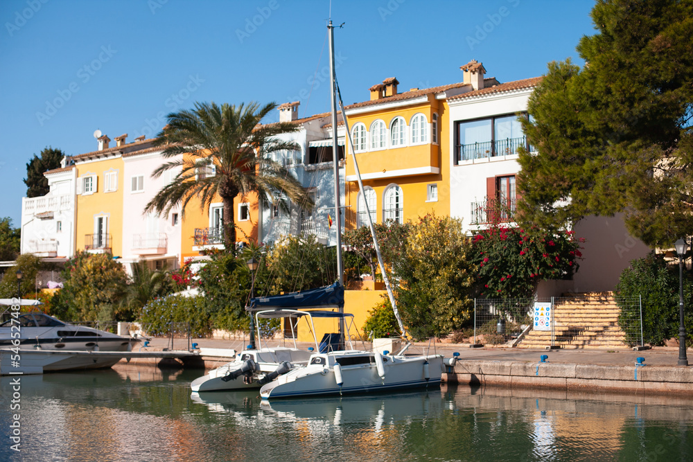 Colorful yellow houses, yachts and palm trees in Port Saplaya, Valencia, Spain on a sunny day landscape. Street and canal with boat in Europe. Travel destination for relaxing vacation.