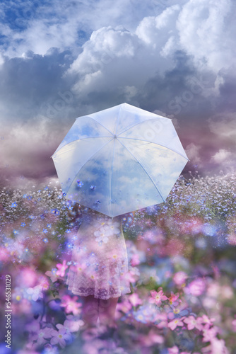 A woman is standing in a flower field with a breeze of blossoms, floating out from an umbrella, that seems to have a blue sky in it, as a metaphor for positivity, hope or belief.