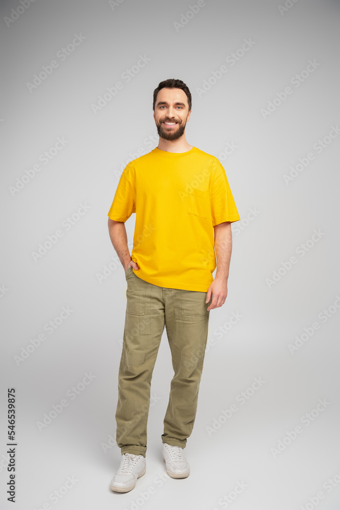 full length of happy bearded man in beige pants and yellow t-shirt standing with hand in pocket on grey background. full length of happy bearded man in beige pants and yellow t-shirt standing with hand in pocket on grey background.