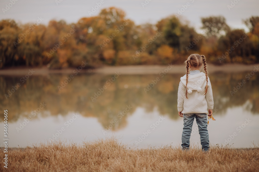 cute little girl stands on the bank of the river in park. autumn, reflections of trees with yellow orange leaves) family leisure. adventure, fun trip with kids. happy travel and vacation concept
