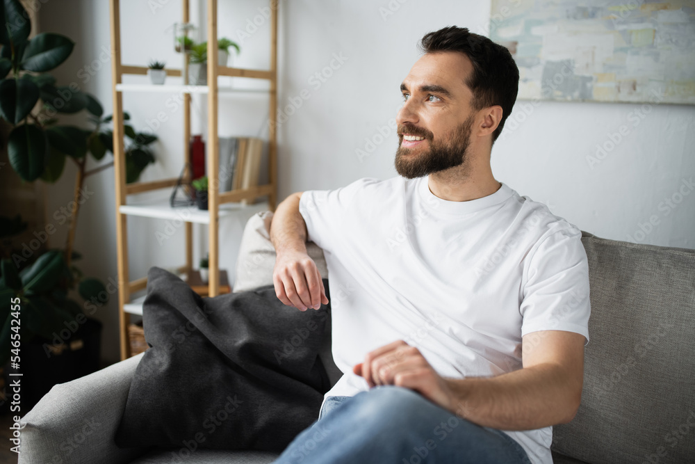 pleased and bearded man in jeans sitting on couch in modern living room.