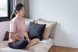 Side view portrait with copy space of cheerful young Asian woman sitting on a couch near windows, crossing legs, practicing yoga meditation, putting hand in hand, closing eyes. Mental health concept