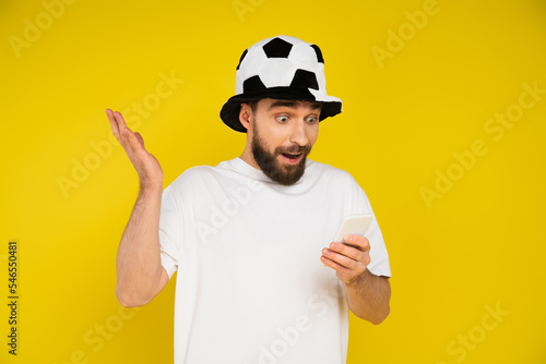 surprised football fan in hat watching game on mobile phone isolated on yellow.